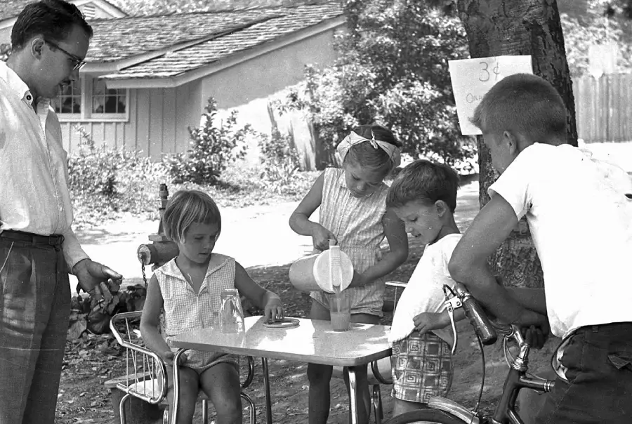 Three children run a lemonade stand, serving drinks to a man and another child on a bicycle outside. A sign reads “3¢”. The scene is set in a shaded, residential neighborhood on a sunny day.