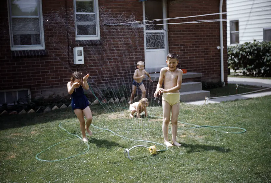 Four young children in swimsuits play in the spray of a lawn sprinkler on green grass in a backyard, with a brick house and a white door in the background.