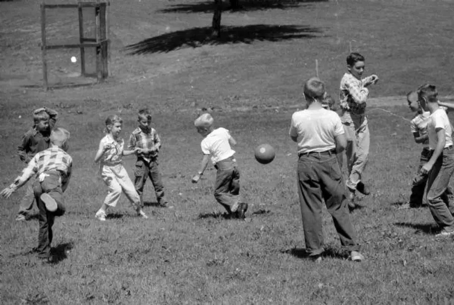 A group of boys play with a ball on a grassy field. Most are mid-action, wearing long pants and plaid shirts. Trees and a wooden structure are visible in the background. The scene appears to be from decades past.