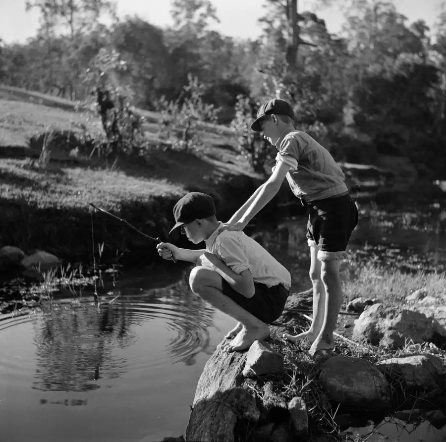 Two boys in shorts and caps, one crouching and fishing while the other stands behind him on rocks by the edge of a pond, surrounded by trees and grass, in a black and white outdoor scene.