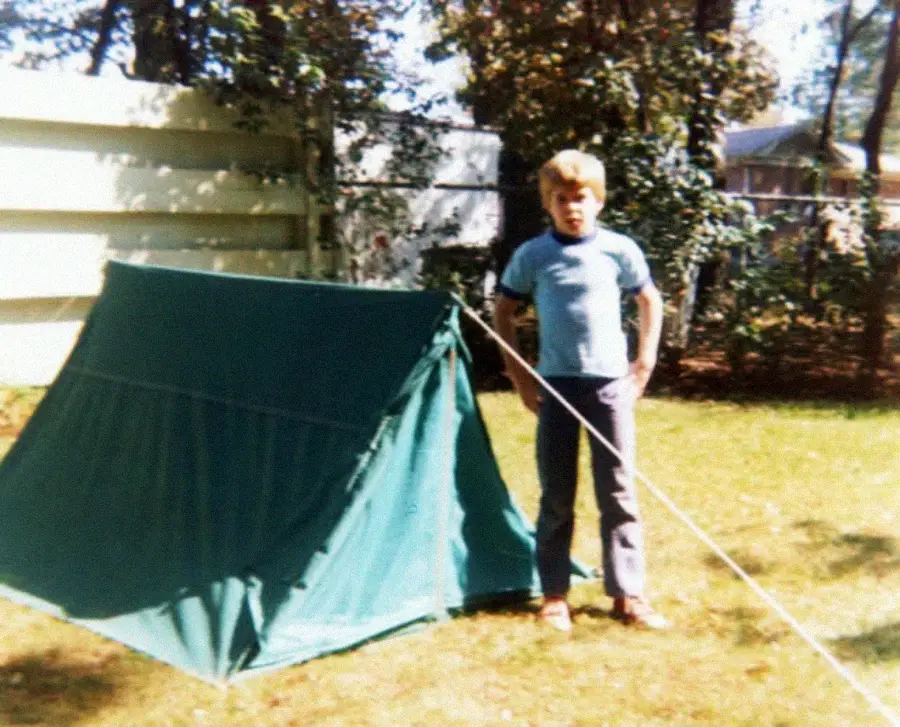 A young boy stands next to a small green tent set up on grass in a sunny backyard, with trees and a wooden fence in the background.