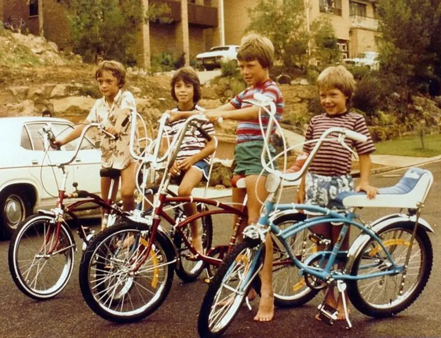 Four children stand on a street with vintage bicycles, smiling at the camera. Houses, trees, and a white car are visible in the background. The kids wear casual summer clothing, and the scene appears to be from the 1970s or 1980s.