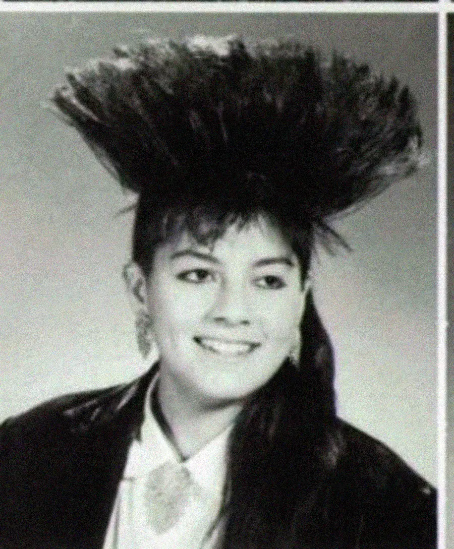 Smiling young woman in a formal jacket and shirt, with large, voluminous hair styled dramatically upward, in a black and white portrait.