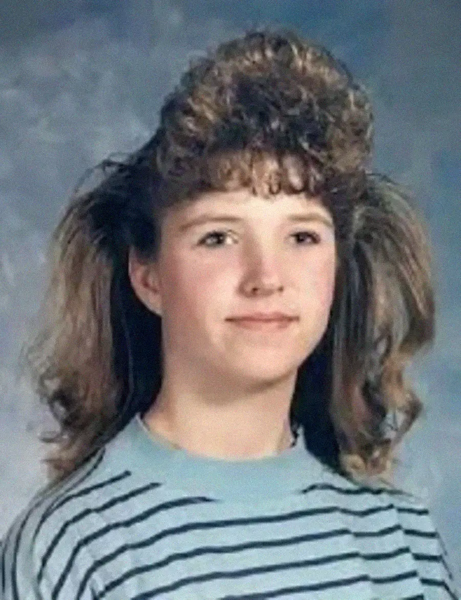 A young person with light skin and voluminous, curly hair styled high on top poses for a school portrait, wearing a light blue and gray striped sweater against a mottled gray background.