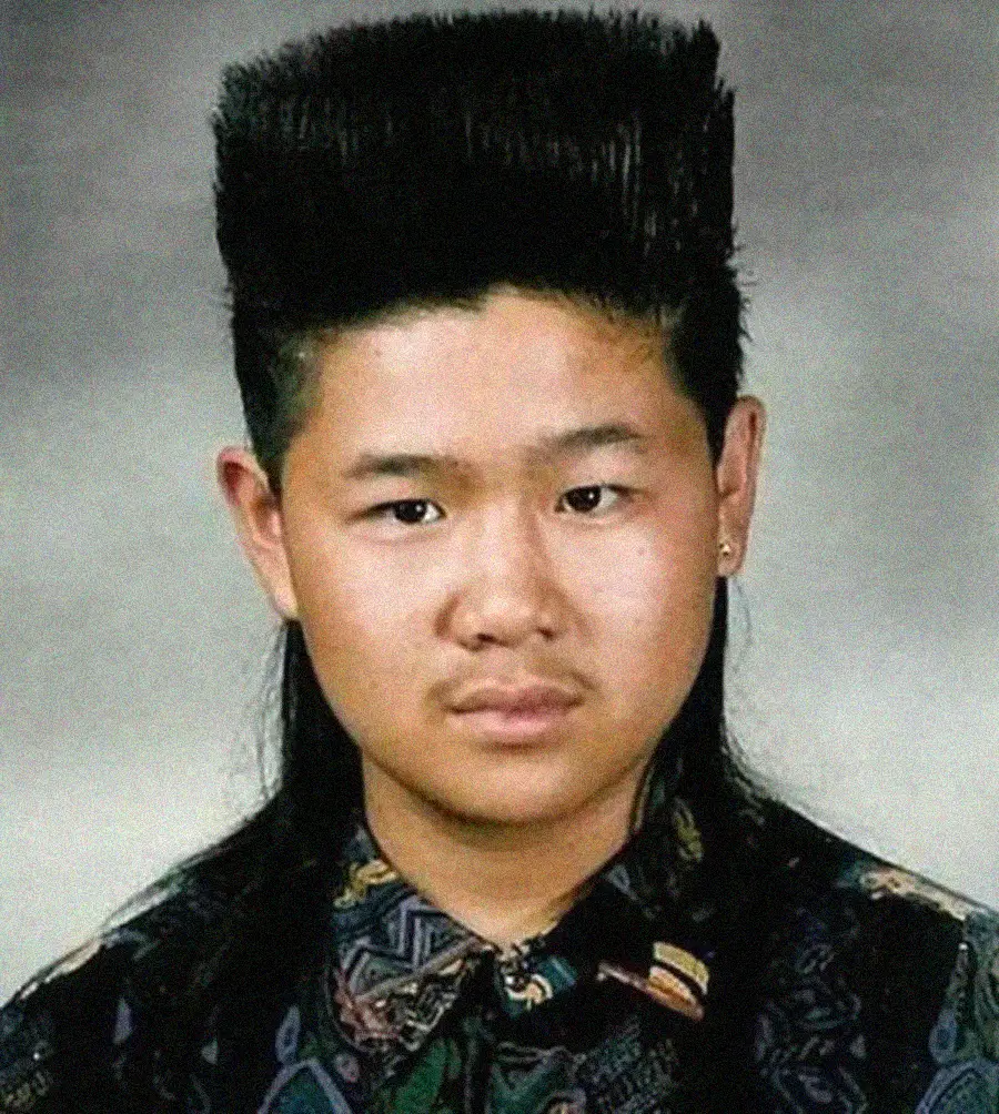 A young person with straight, black hair styled in a tall flat top mullet poses for a school portrait, wearing a patterned button-up shirt and a small earring, against a neutral gray background.