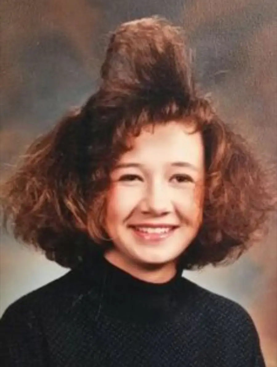 A young person with curly brown hair styled in a tall, teased beehive shape smiles while wearing a dark sweater, posing for a school portrait against a neutral background.