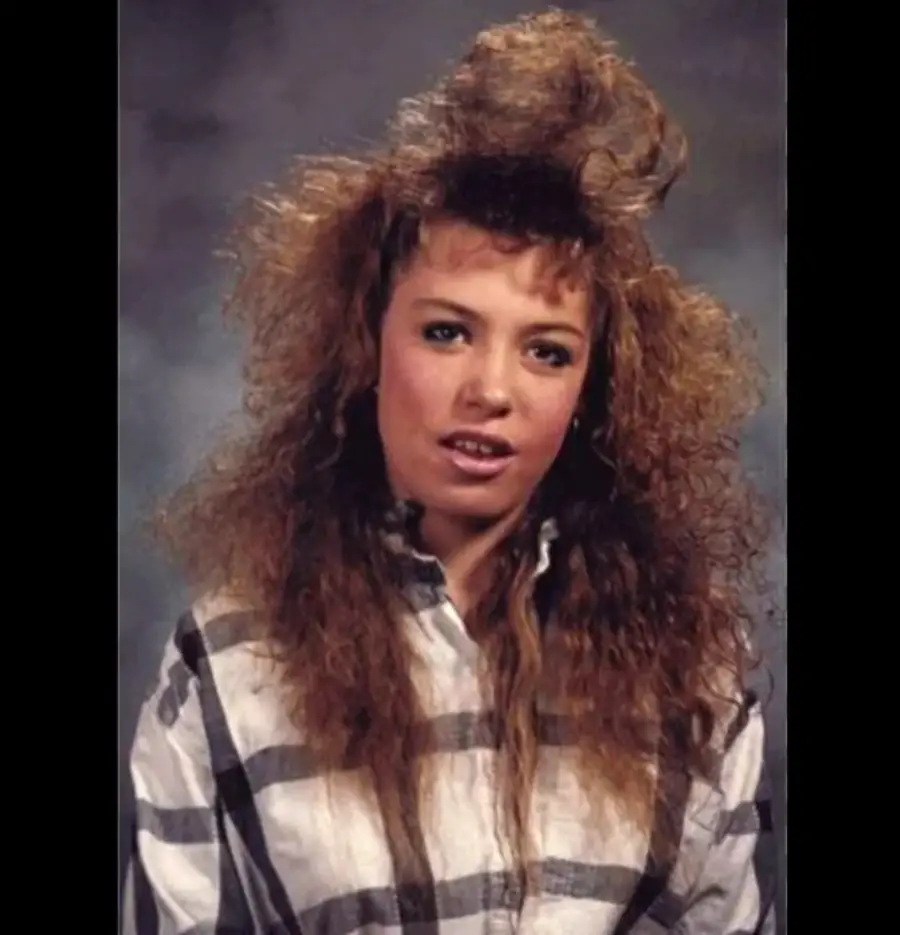 A young woman wearing a plaid shirt poses for a portrait with voluminous, teased curly hair styled in a dramatic 1980s fashion against a gray studio background.