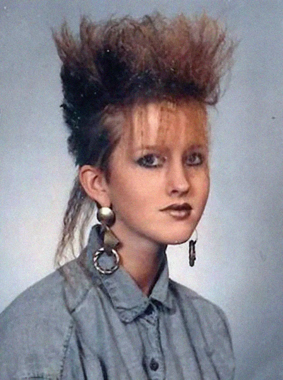 A young woman poses for a portrait wearing a gray button-up shirt, large statement earrings, and dramatic teased hair styled high on top. Her makeup is bold, with dark eyeliner and lipstick.