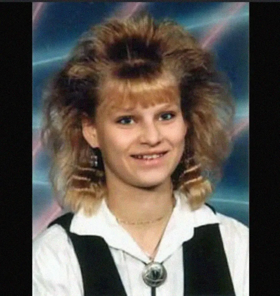 A young woman with voluminous blonde hair and bangs smiles, wearing a white shirt, black vest, and a bolo tie.