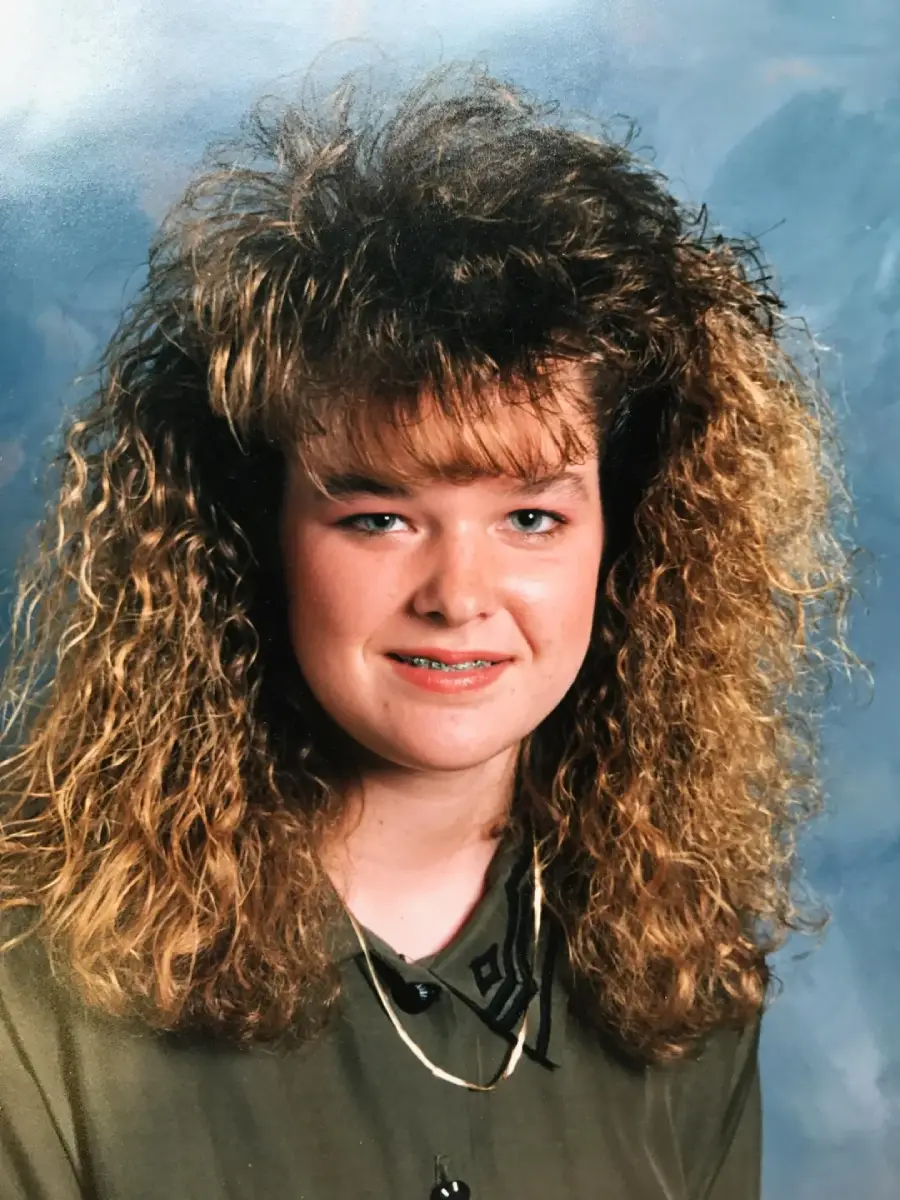 A young woman with voluminous, curly hair styled in an 80s fashion, wearing a green shirt and a gold necklace, smiles at the camera against a blue-gray studio backdrop.