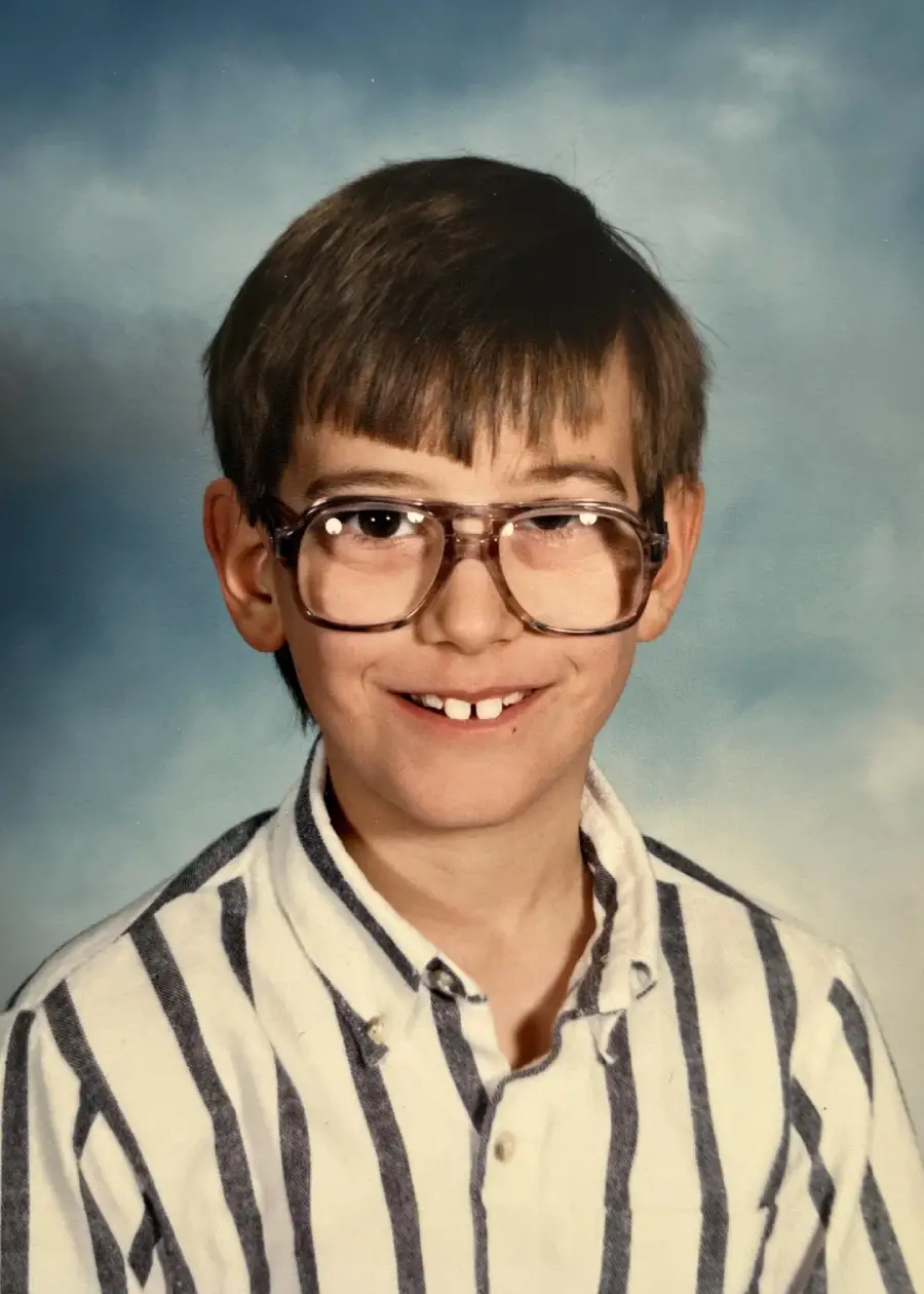 A young boy with light brown hair and large glasses smiles at the camera. He is wearing a white shirt with bold, dark vertical stripes. The background is a simple blue and gray gradient, typical of school portraits.