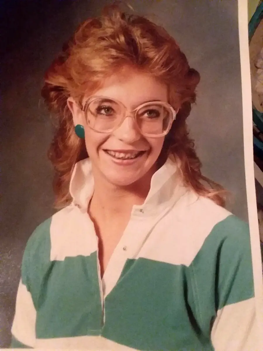 A young woman with wavy, reddish hair wears large, round glasses, teal earrings, and a green-and-white striped collared shirt, smiling against a studio portrait backdrop.