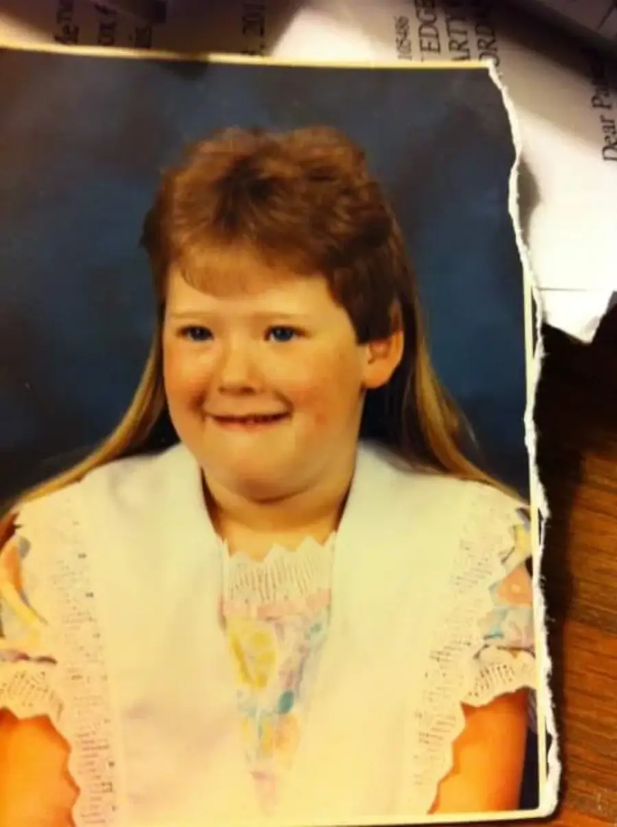 A school portrait of a young girl with strawberry-blonde hair in a mullet hairstyle, wearing a white vest over a pastel floral dress, smiling in front of a blue background. The photo’s edge is slightly torn.