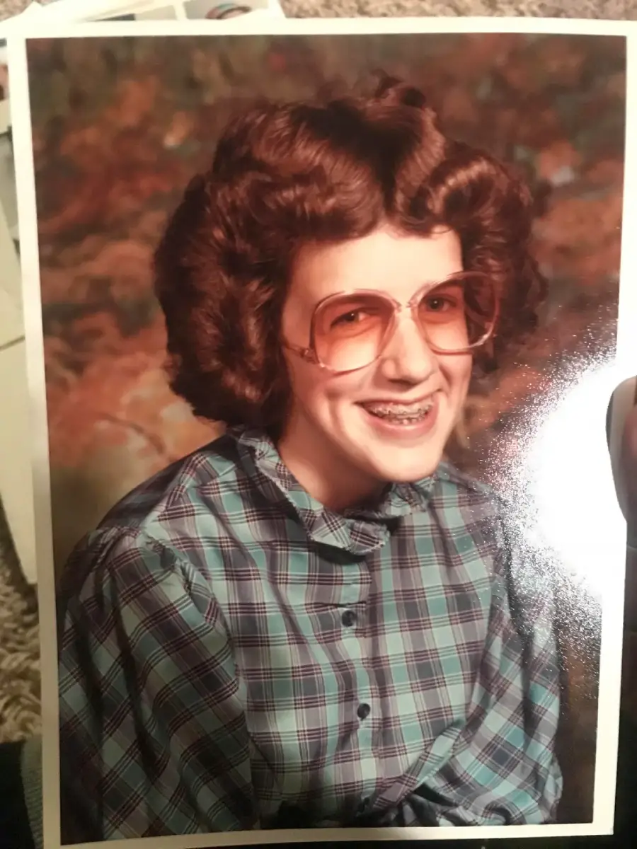 A person with curly brown hair, large tinted glasses, and braces smiles for a portrait. They are wearing a blue and purple plaid shirt with a high collar. The background is a mottled, warm-toned studio backdrop.