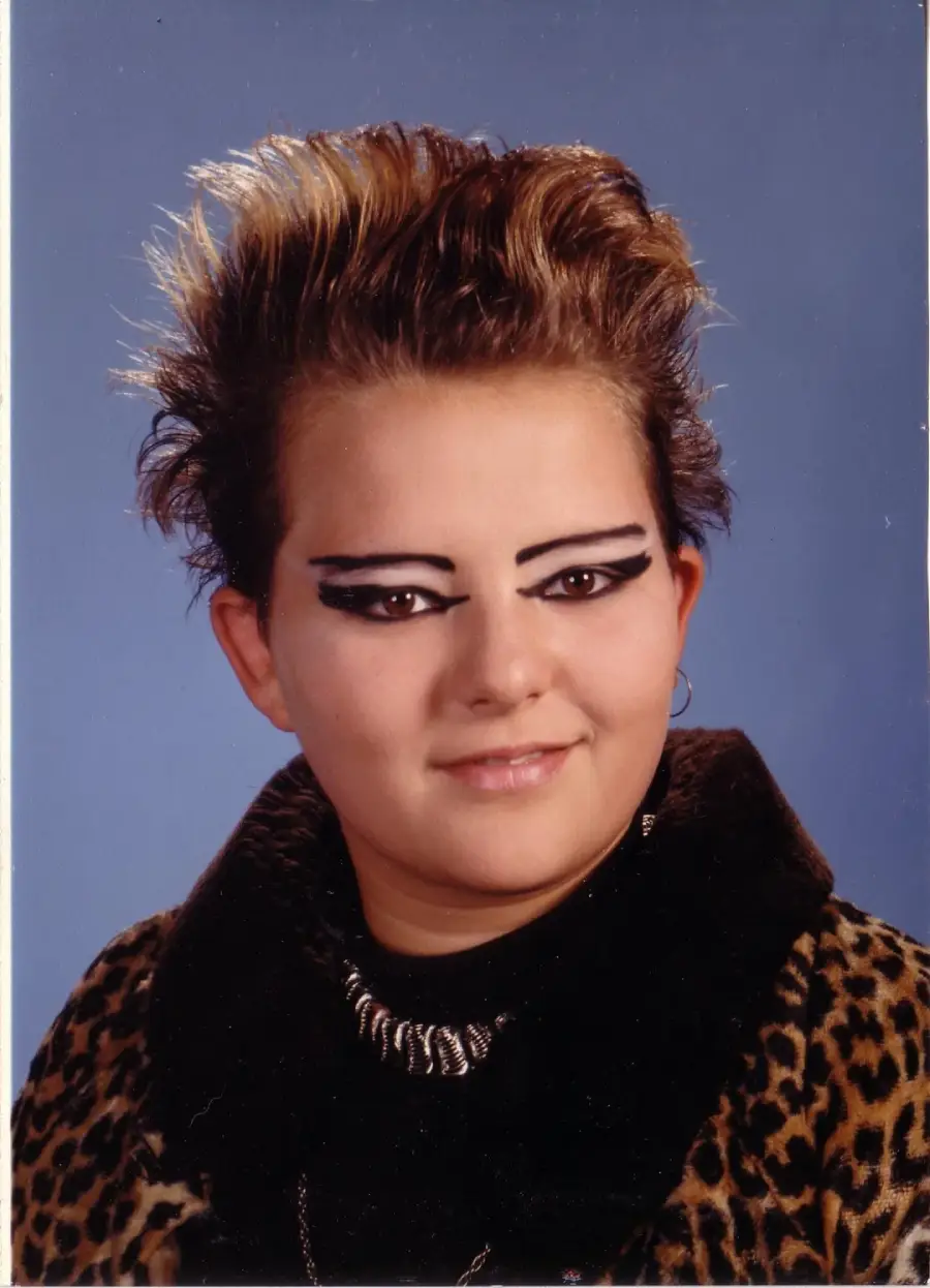 A person with short, spiked hair and dramatic black eyeliner poses for a portrait against a blue background, wearing a leopard-print coat and a chunky metallic necklace.