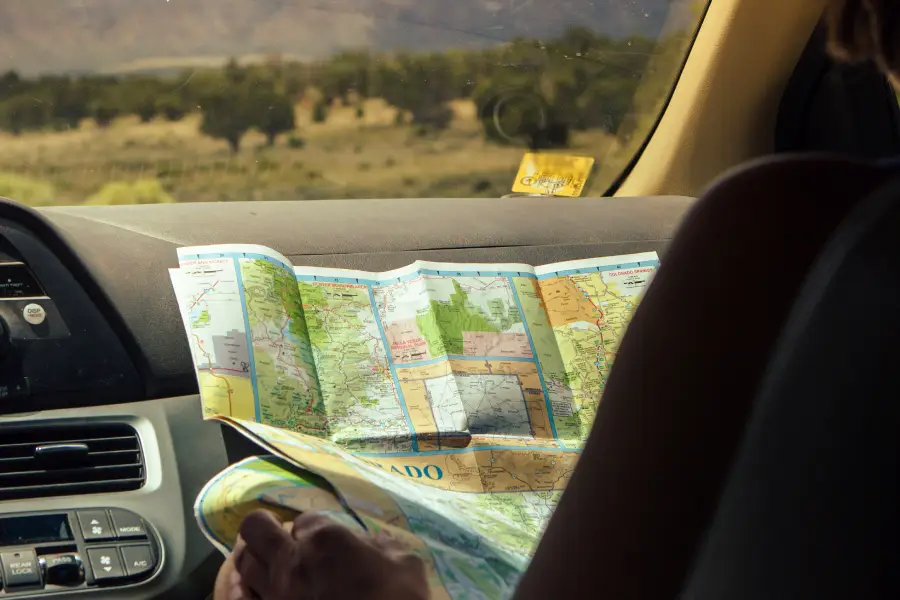 A person sits in a car holding and studying a large, unfolded map. The car dashboard and part of the steering wheel are visible, with an outdoor landscape and trees seen through the windshield.