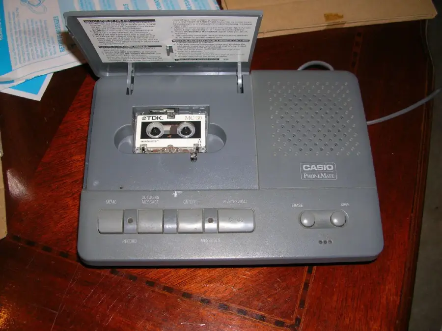 A gray Casio PhoneMate answering machine with an open cassette tape compartment, showing a TDK mini tape inside. The machine has several buttons and a speaker on the front, and sits on a wooden surface.