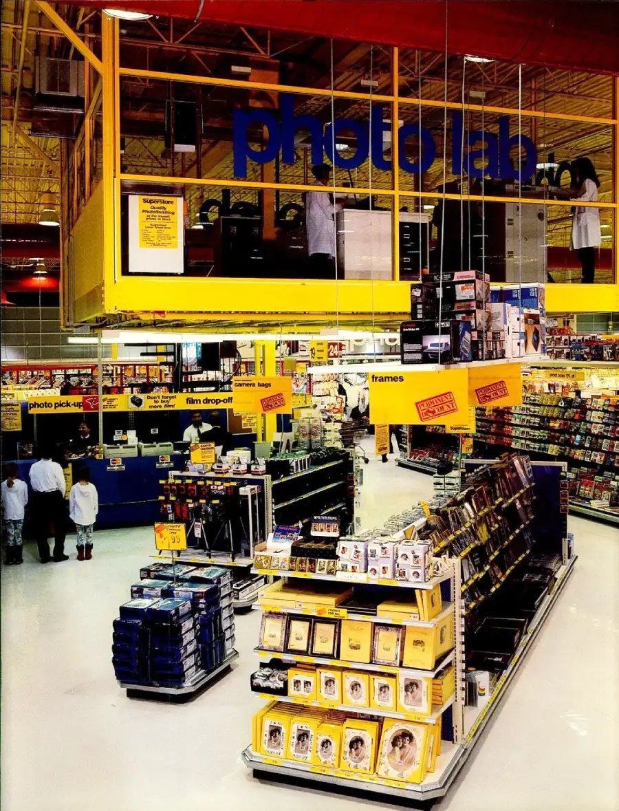 A brightly lit store interior with a large yellow "photo lab" section above rows of photo frames and film products. Shoppers browse aisles filled with photo-related items and other store products.