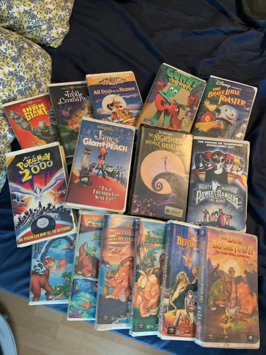 A collection of VHS tapes featuring classic animated movies like The Lion King, Pokémon 2000, James and the Giant Peach, The Nightmare Before Christmas, and others, laid out on a blue bedspread.