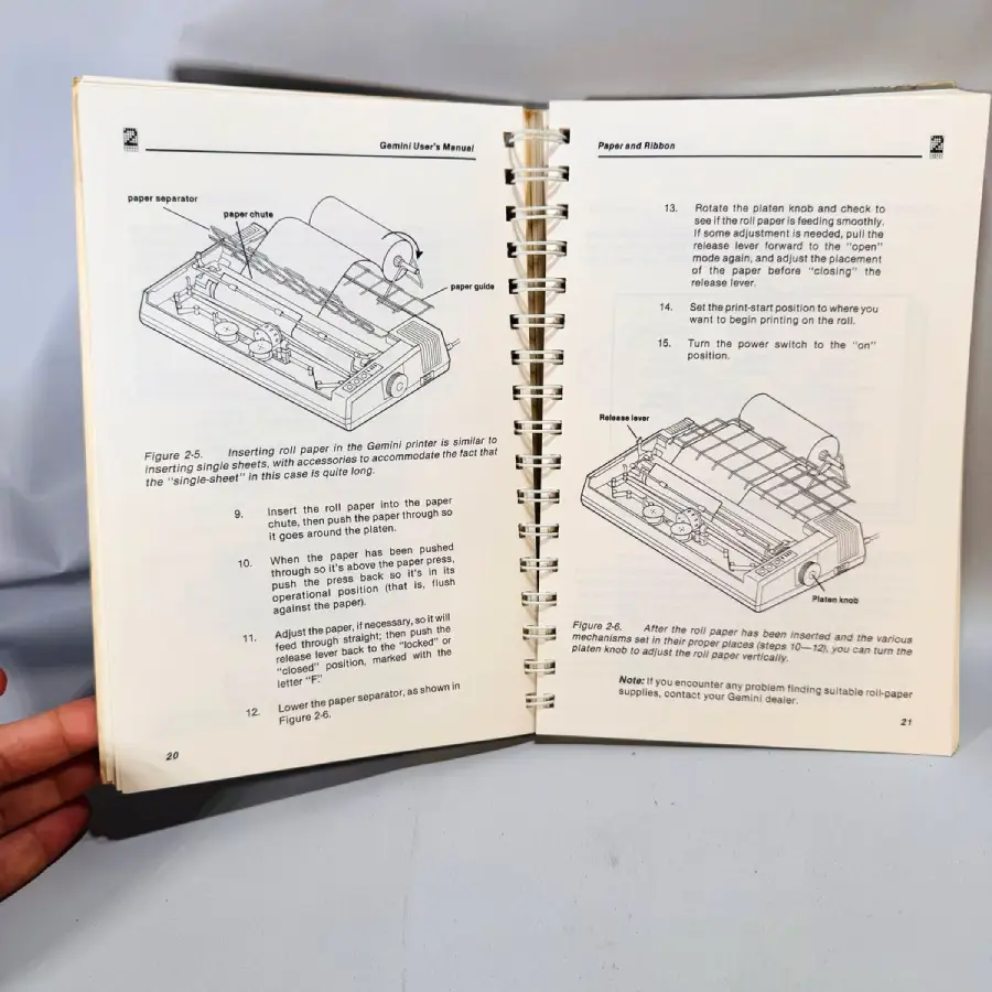 An open manual showing diagrams and step-by-step instructions for loading paper into a printer. The pages include labeled illustrations of printer parts and numbered text directions for proper setup. A hand holds the left side.
