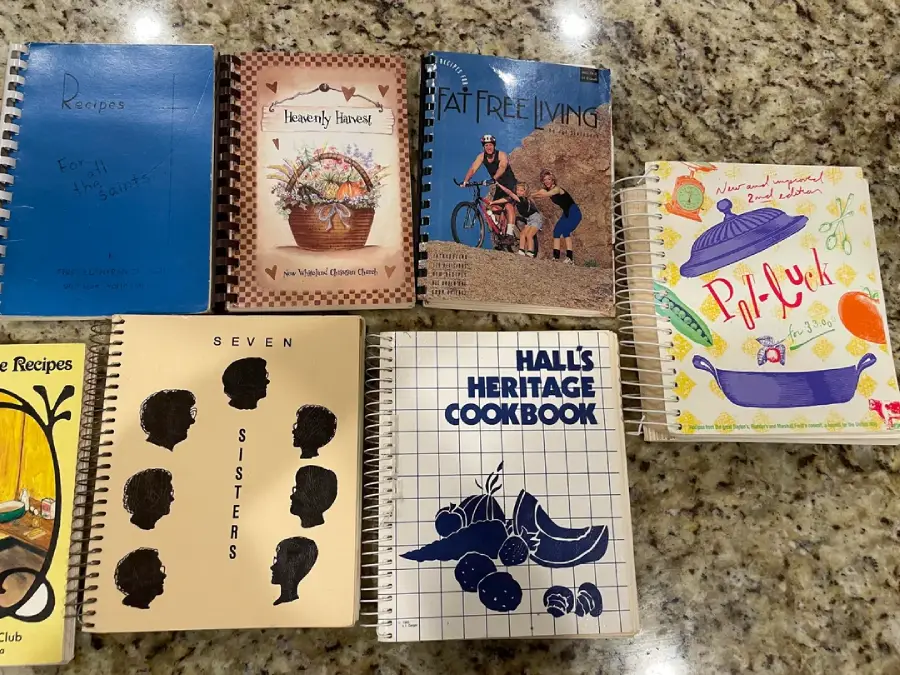 Seven spiral-bound community cookbooks are arranged on a granite counter, each with colorful and unique covers featuring food illustrations, silhouettes, and titles relating to family, heritage, and group recipes.