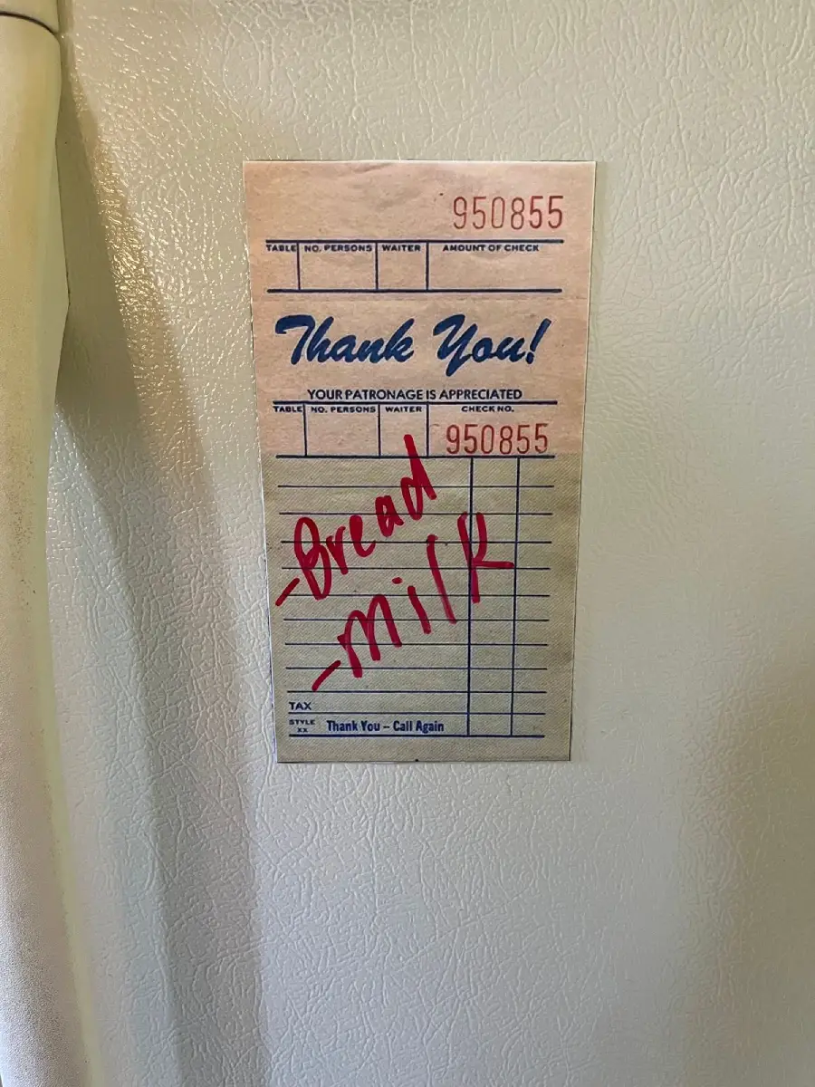 A restaurant receipt stuck to a fridge with the words "Bread" and "Milk" written in red marker across it, serving as a grocery reminder.