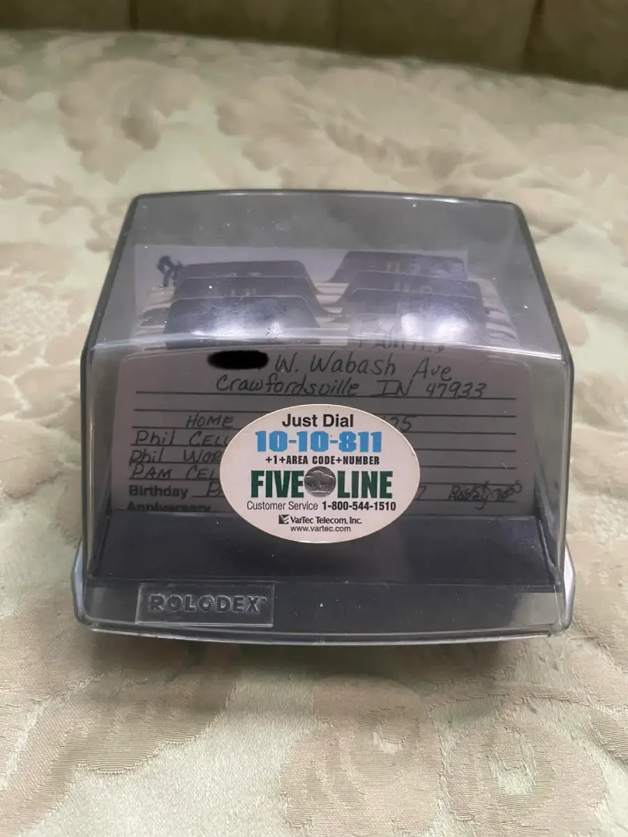 A Rolodex with contact cards inside sits on a beige patterned surface. A sticker on the front displays dialing instructions for 10-10-811 long-distance service and customer support information. Handwritten addresses are visible inside.