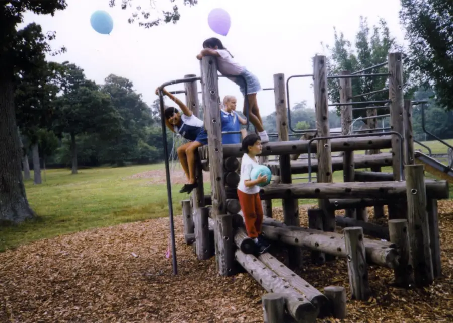 Four children play on a wooden playground structure in a park. Two kids climb a pole, one stands on the structure, and another holds a ball on the ground. Balloons are visible in the background among trees.