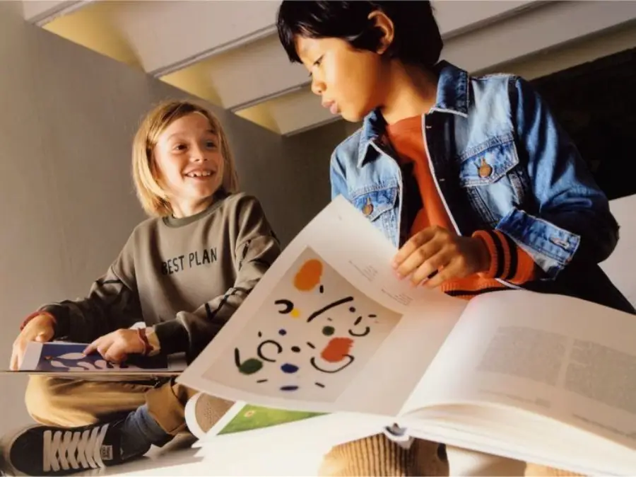 Two children sit side by side, smiling and looking at each other while holding open books. One child wears a denim jacket and the other a green sweatshirt that reads "BEST PLAN." The books have colorful abstract art inside.
