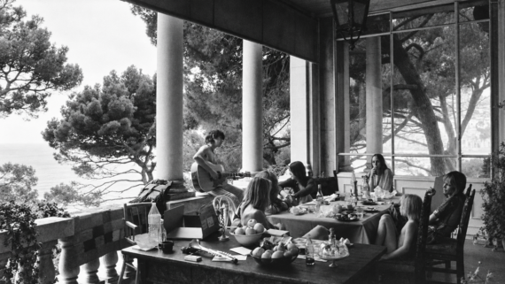 A group of people sitting at a table on a shaded veranda overlooking the sea, with one person playing an acoustic guitar.