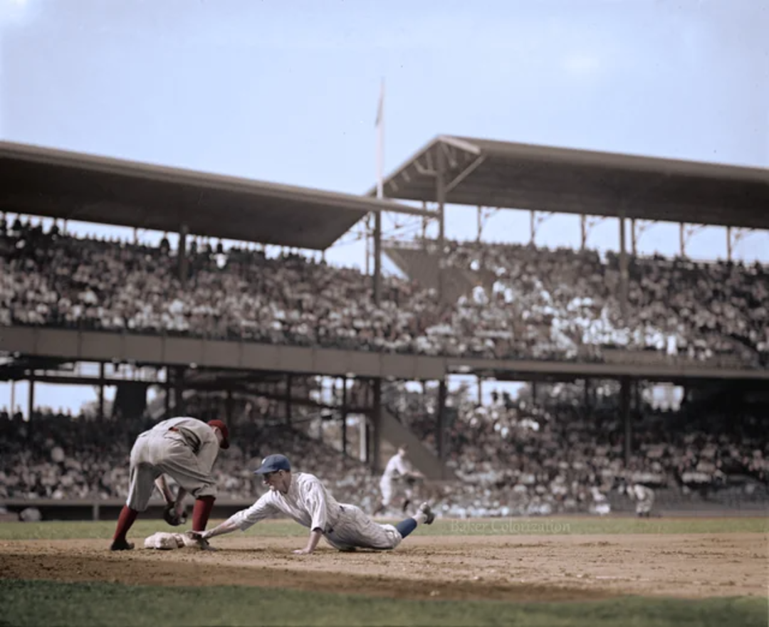 A baseball player in a white uniform slides into a base as an opposing player in a gray and red uniform tries to tag him, with a packed stadium in the background.