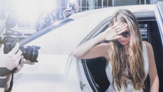 A woman with long hair and sunglasses steps out of a white car, shielding her face from bright camera flashes as photographers take pictures outside.