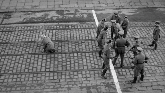 A group of uniformed soldiers stand and talk on cobblestone tram tracks, while one person kneels alone nearby. The scene appears tense and is viewed from above.