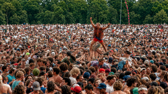 A large crowd at an outdoor event with a person crowd-surfing above, raising one arm in the air. The scene is lively, with dense groups of people surrounded by trees in the background.