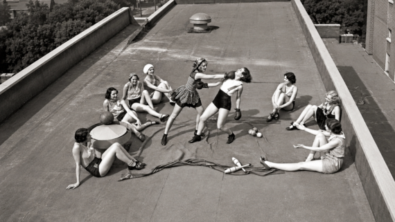 A group of women in vintage swimsuits sit on a rooftop, watching two women playfully boxing while others relax around them.