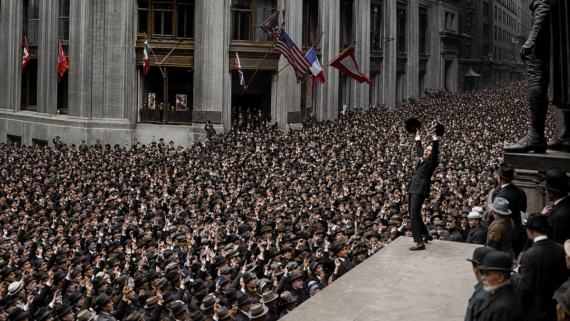 A man stands on a platform addressing a dense crowd of people in suits and hats outside large buildings decorated with flags.