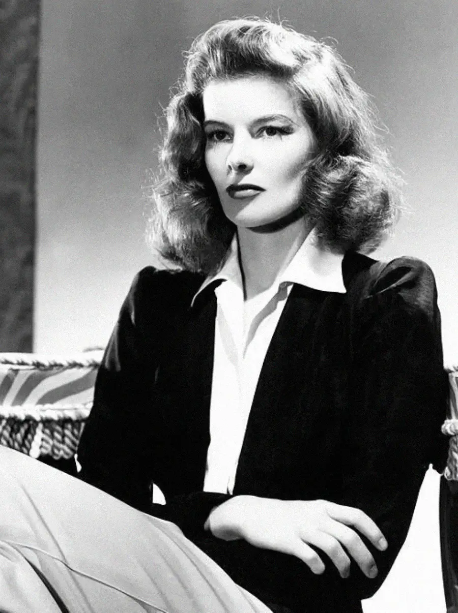 Black-and-white portrait of a woman with wavy, shoulder-length hair, wearing a dark jacket over a light blouse, sitting with arms folded and looking slightly to the side with a serious expression.