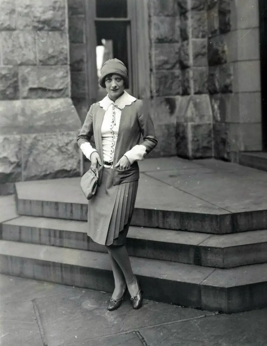 A woman in 1920s fashion stands on stone steps in front of a building, wearing a cloche hat, pleated skirt, jacket, and holding a small purse, looking at the camera with a neutral expression.