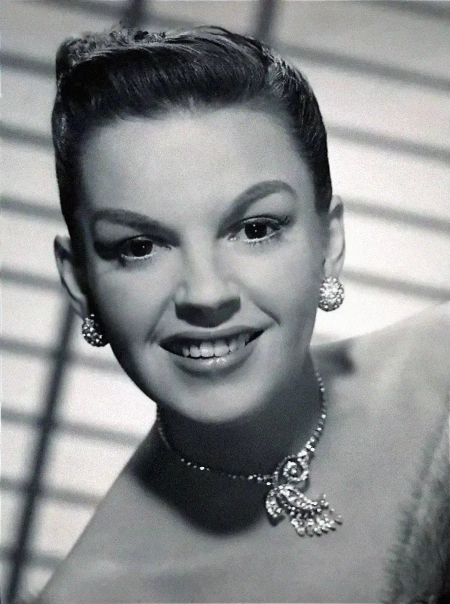 A black and white portrait of a woman smiling, wearing ornate earrings and a jeweled necklace, with her hair styled up. Horizontal shadows in the background add contrast to the image.