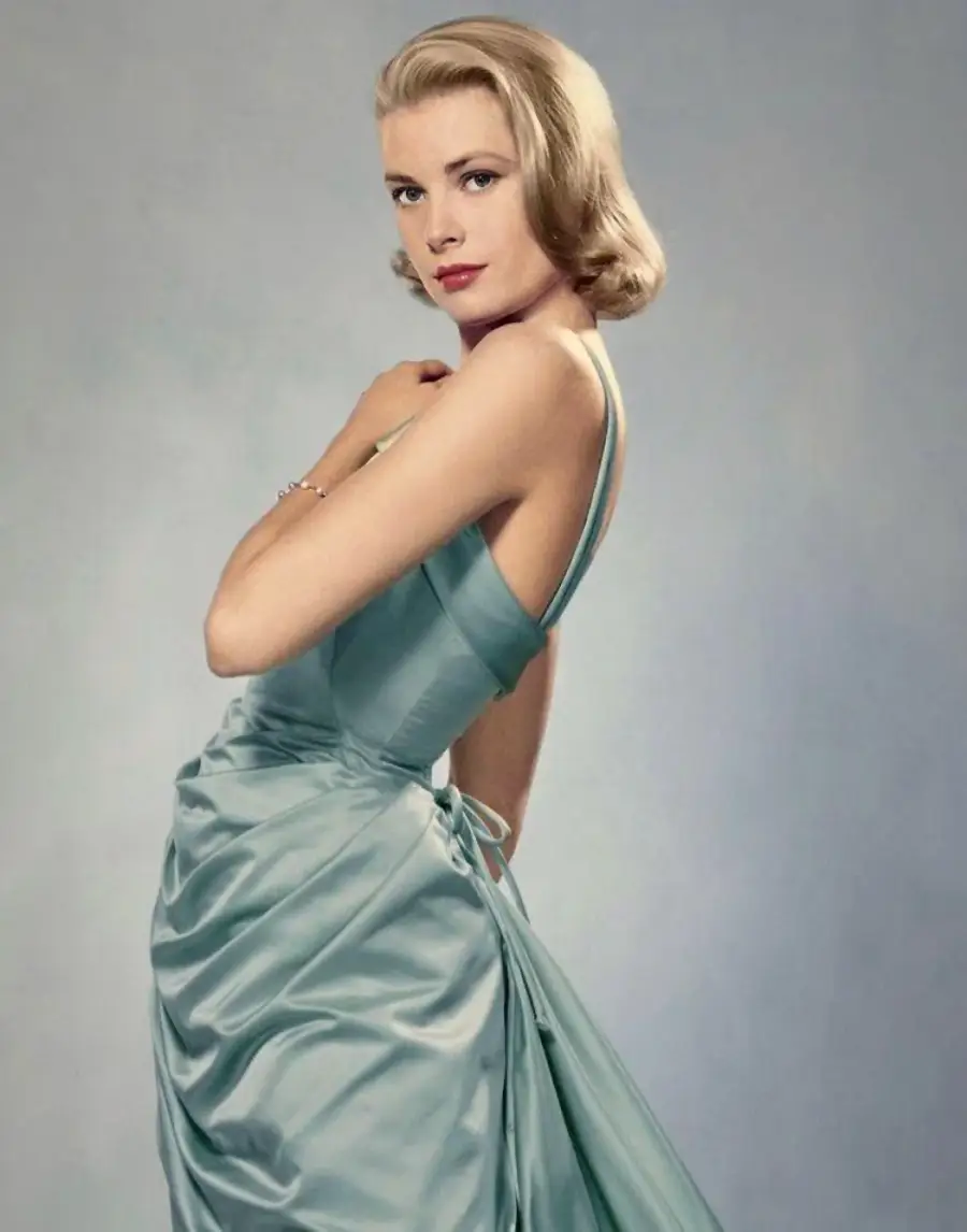 A woman with short blonde hair wears a light blue satin gown with a fitted bodice and full skirt, posing gracefully against a plain, light-colored background. She looks over her shoulder with a soft expression.