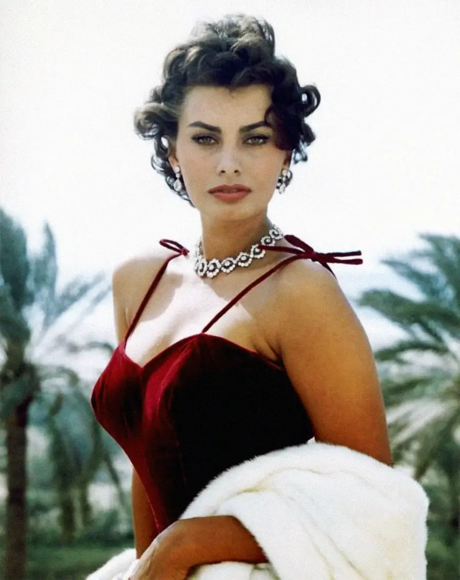 A woman with short, curly dark hair and striking makeup wears a red velvet dress, a diamond necklace, and earrings. She holds a white fur stole, standing outdoors with blurred palm trees in the background.