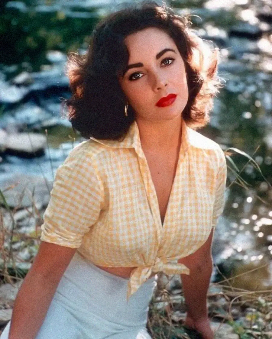 A woman with wavy dark hair and bright red lipstick sits outdoors by a sunlit stream. She wears a yellow gingham blouse tied at the waist and a light-colored skirt, gazing at the camera with a calm expression.