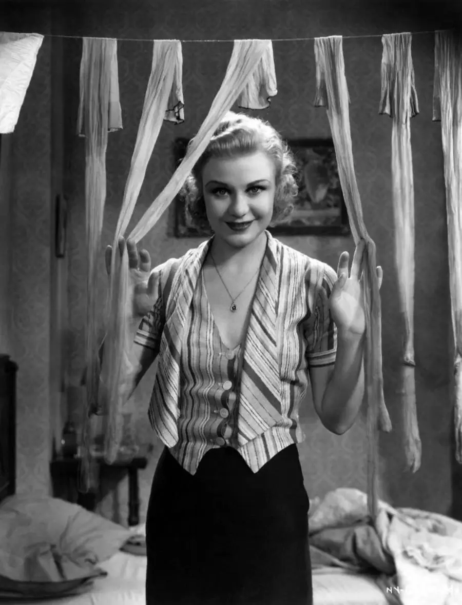 A woman with wavy hair stands smiling in a room, parting hanging strips of cloth that frame her face. She wears a striped blouse and a pendant necklace, and the background includes a bed and a patterned wall.