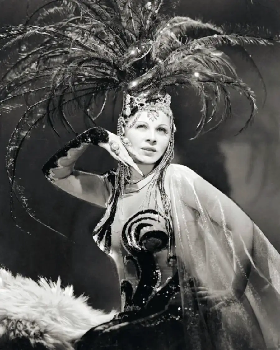 A woman in a glamorous costume poses with her hand on her chin, wearing a sparkling headpiece adorned with large feathers and a sequined dress with sheer, flowing sleeves. The image is in black and white.