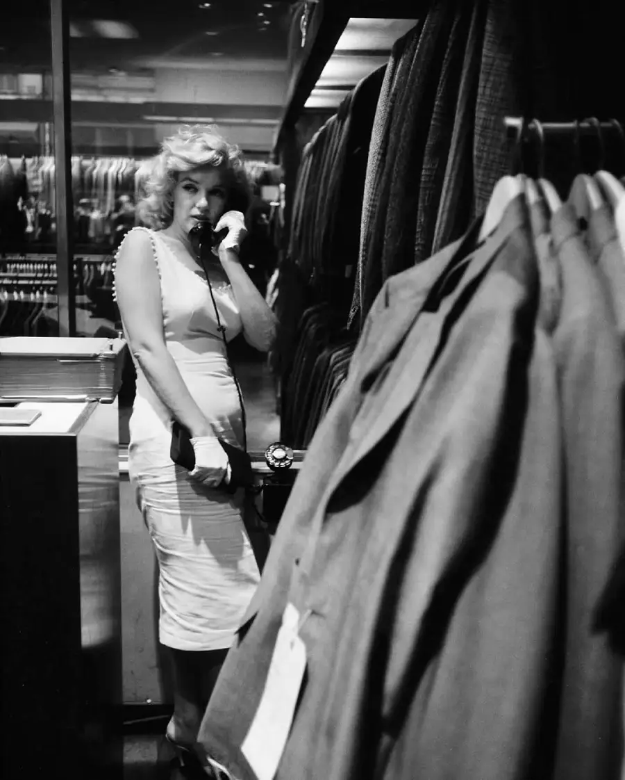 A woman in a light dress and gloves stands in a clothing store, speaking on a telephone. She is surrounded by racks of suits and appears to be looking toward the camera. The photo is black and white.