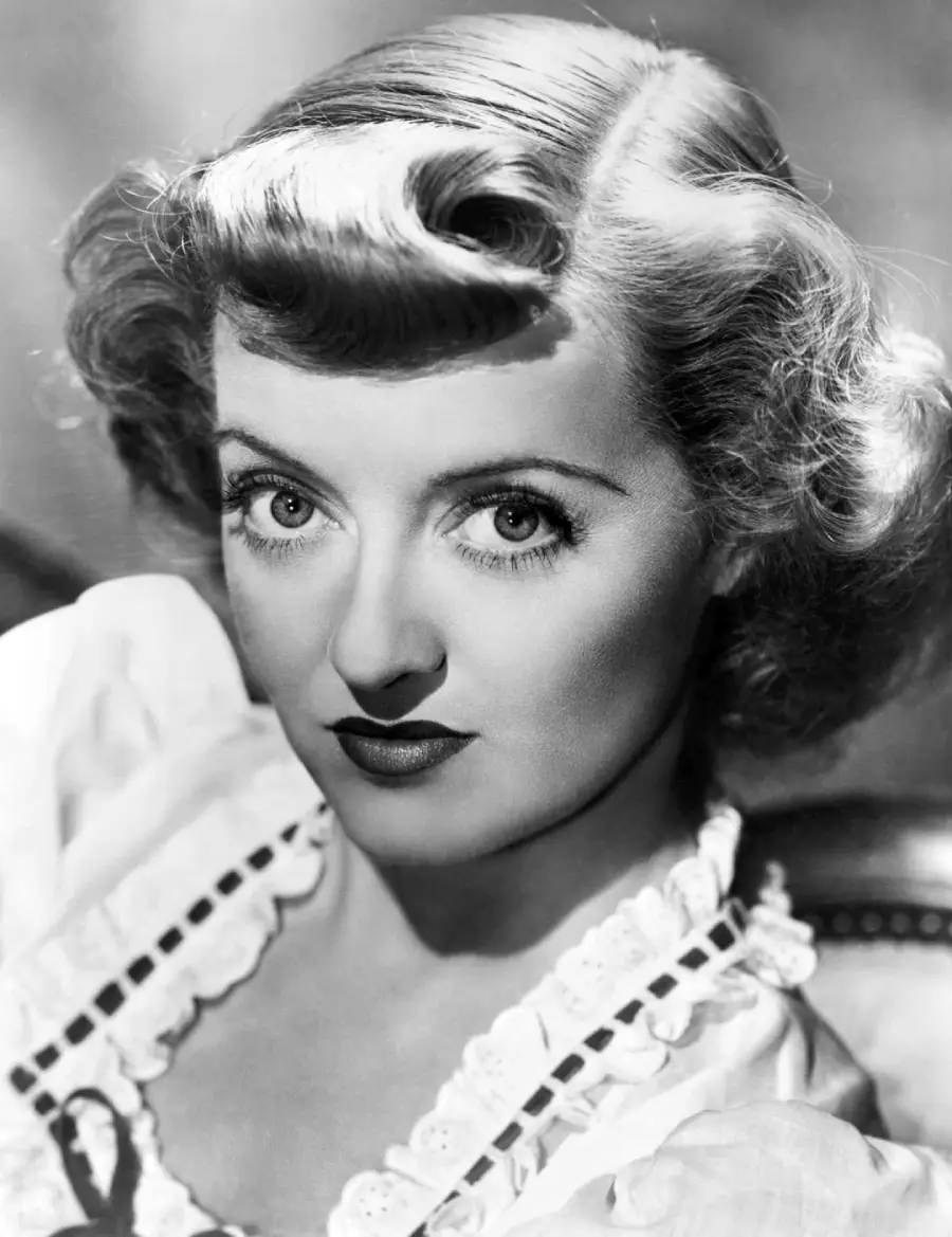 Black and white portrait of a woman with wavy, shoulder-length hair, styled in retro waves. She has arched eyebrows, dark lipstick, and is wearing a blouse with lace trim, looking directly at the camera.