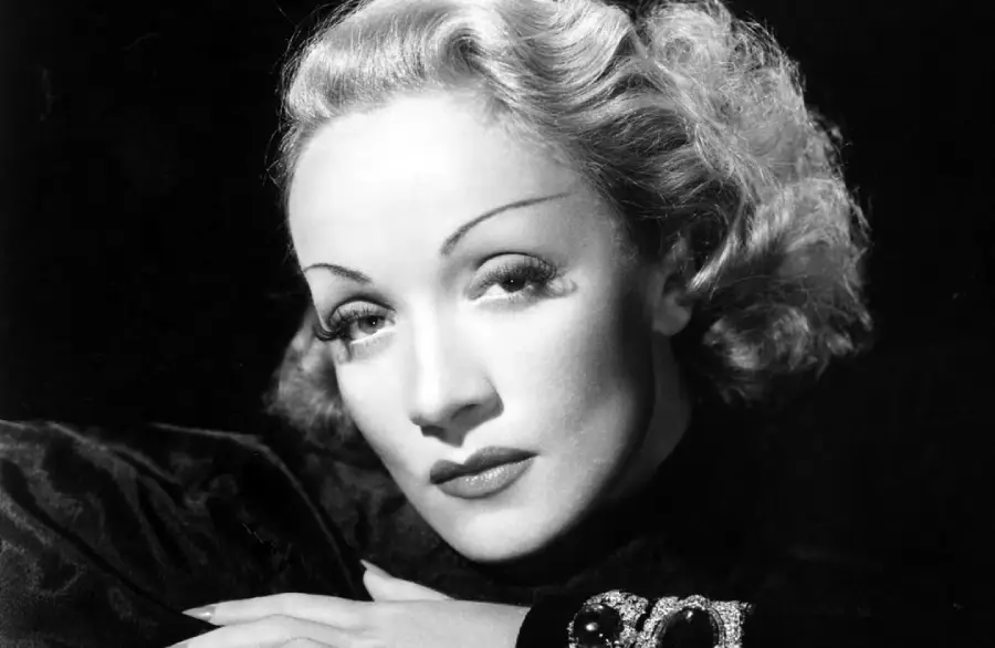 Black and white portrait of a woman with wavy, blonde hair, thin arched eyebrows, and dramatic eye makeup, resting her hands on a surface and gazing softly at the camera.