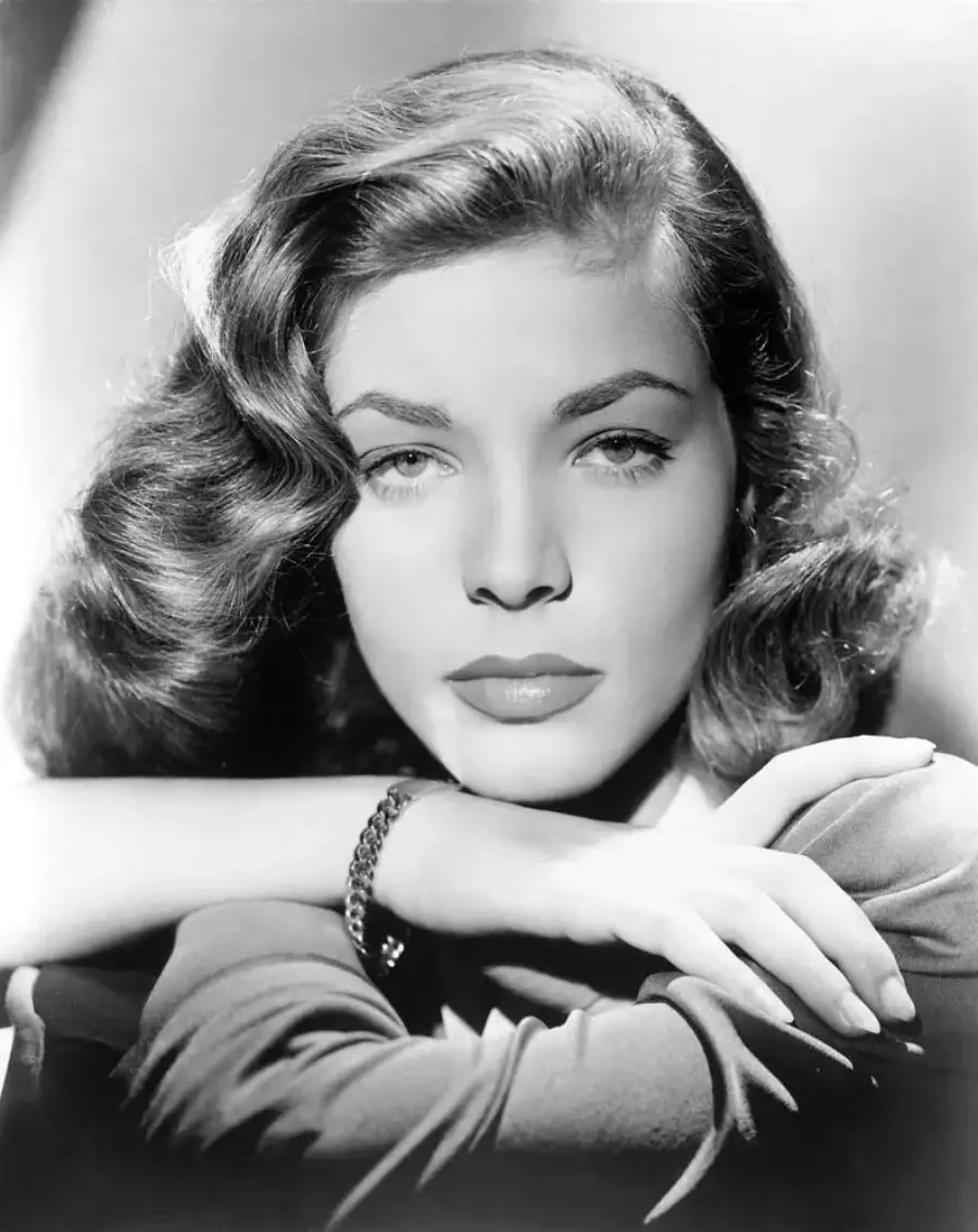 Black and white portrait of a woman with wavy hair, resting her chin on her crossed arms, looking directly at the camera with a neutral expression. She wears a bracelet and has classic 1940s-style makeup.