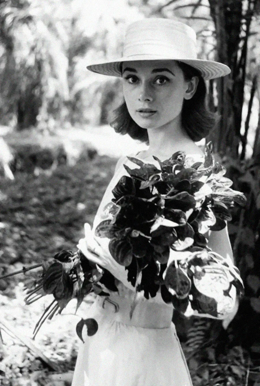 A young woman in a white dress and wide-brimmed hat holds a large bouquet of leafy branches while standing outdoors in a sunlit, wooded area. The black-and-white photo has a classic, vintage feel.