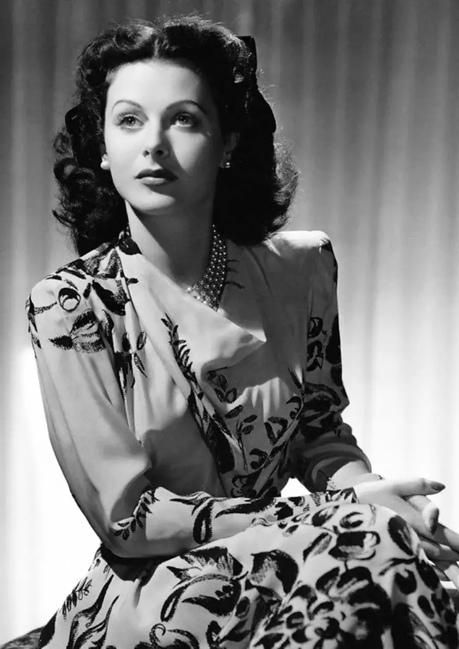 Black and white portrait of a woman with wavy dark hair, wearing a patterned dress and a pearl necklace, sitting with her hands clasped, looking slightly upward. The background has vertical soft lighting.